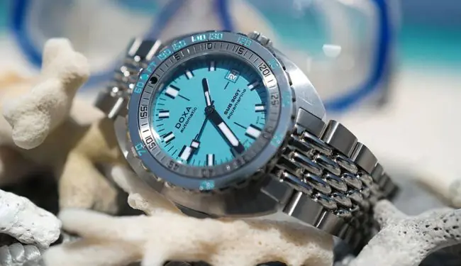 DOXA dive-style watch with a blue dial on a metal bracelet resting on sand and shells